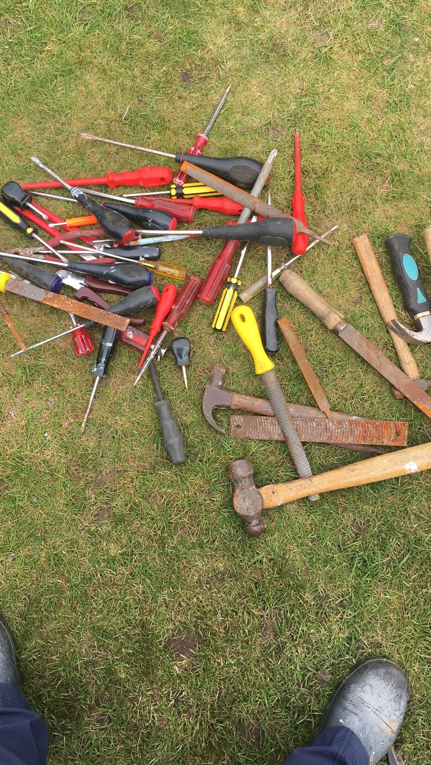 Pile of assorted hand tools (screwdrivers, hammers, files, chisels) scattered on grass with a pair of boots visible nearby.