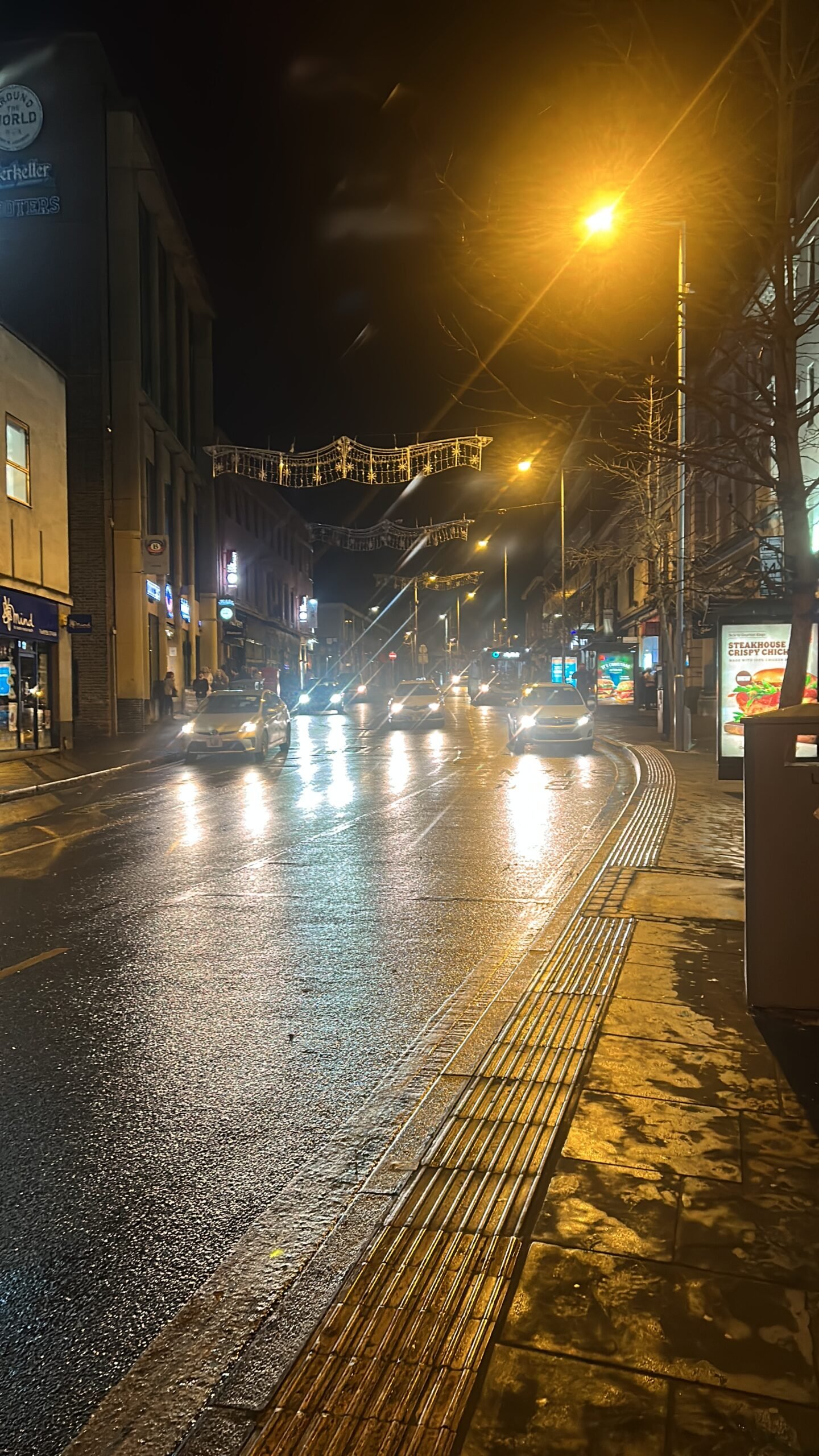 A wet UK high street at night, with street lights and car headlights reflecting off the road.