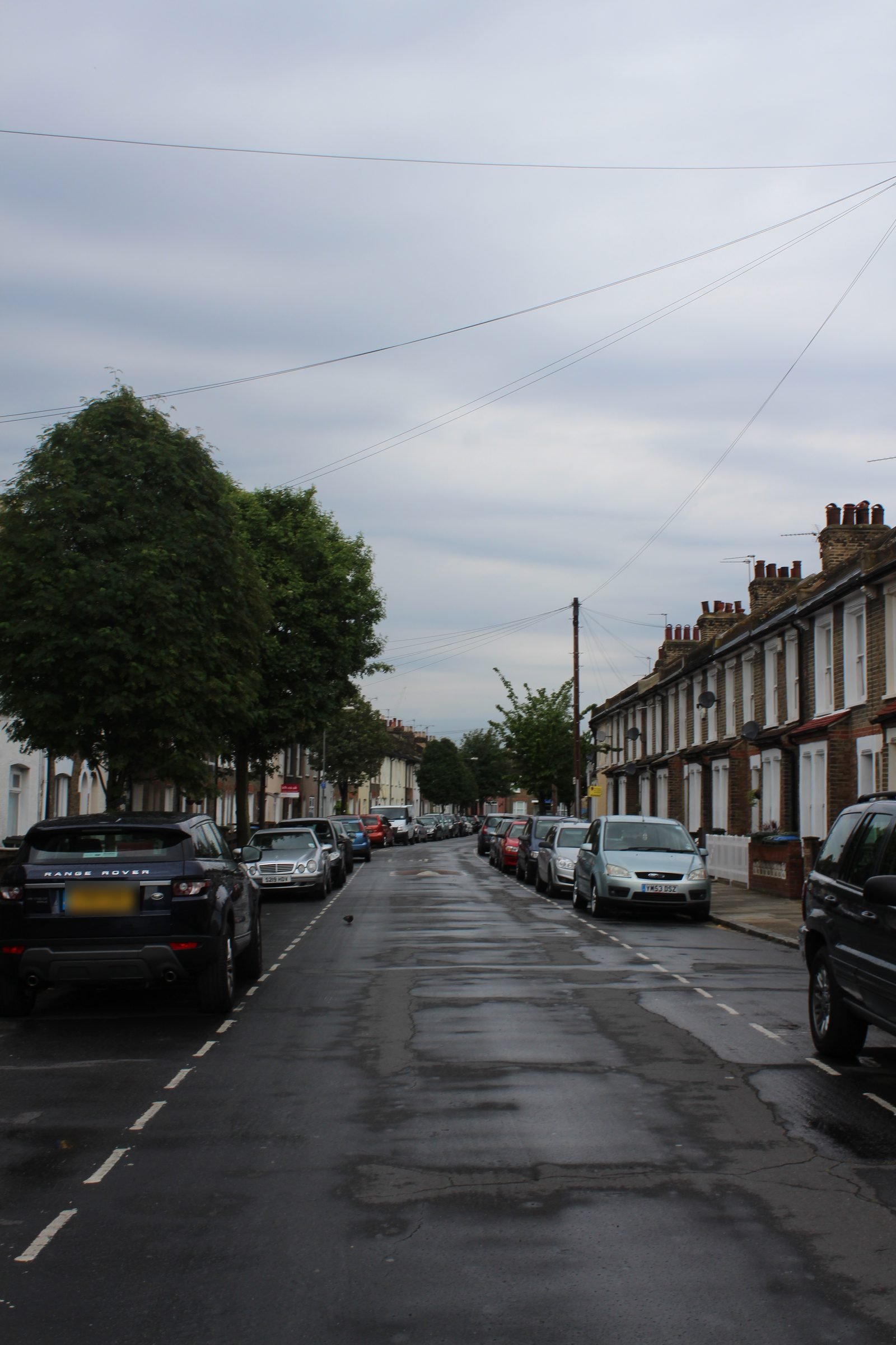 An ordinary UK terraced street with parked cars, trees and a wet road under an overcast sky.